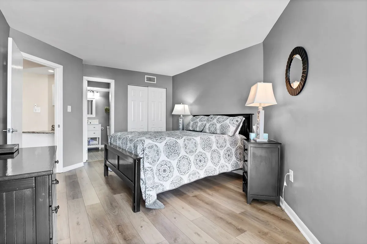 A stylish queen bedroom with a plush bed, nightstand and lamp, mirror, and a doorway leading to a private bathroom. The room is softly lit by natural light, exuding a peaceful and serene atmosphere ideal for relaxation or reading.