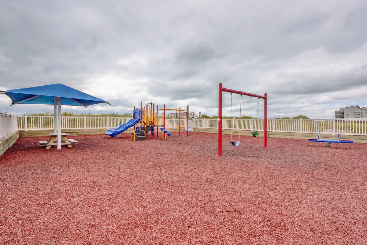 A bright, playful playground complete with swings, climbing equipment, and a picnic table beneath a shade structure, set outdoors and ready for children to enjoy. The setup is clearly designed for family-friendly fun and community gatherings.