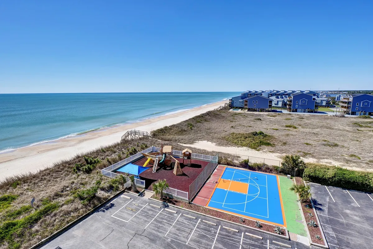 The image showcases the vibrant exterior amenities at the St Regis 1505 condo complex, featuring a colorful playground, full basketball court, and a sweeping view of North Topsail Beach with the ocean bathed in the warm glow of sunset. Children are playing on the playground while families stroll along the sand, capturing a lively yet relaxed atmosphere.