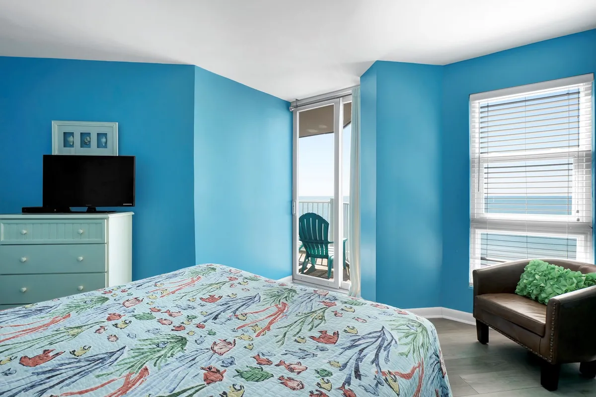 This image features a bed with a floral quilt, a television mounted on the wall, a chair with a green cushion, and a beautiful balcony view of the ocean beyond. Natural light fills the space, creating a bright and inviting mood. The sliding glass doors lead directly onto the balcony, blending indoor comfort with the appeal of coastal scenery.