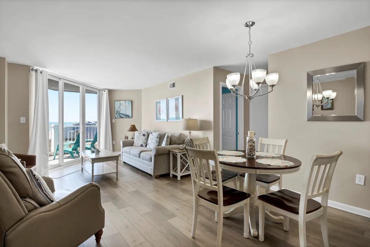 A cozy living area with a sofa and dining table basking in natural sunlight, thanks to sliding glass doors that open up to ocean scenery. The chandelier and decor create a warm, welcoming vibe perfect for family meals or relaxing evenings.