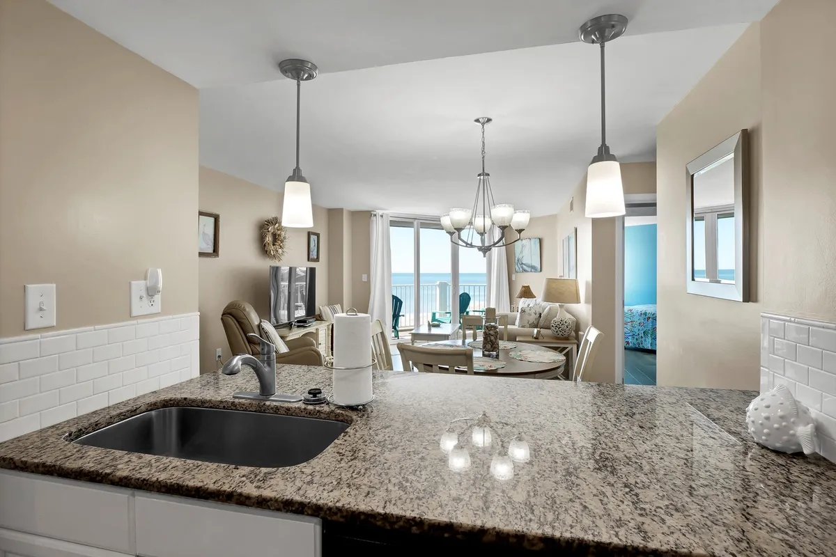 This image captures an inviting open-concept space where both the dining area and living room are visible. Through the large windows, there's a sweeping view of the ocean, filling the room with light. The granite countertop and chandelier bring elegance, while the arrangement of the dining table and living room seating signals flexibility and flow for social gatherings.