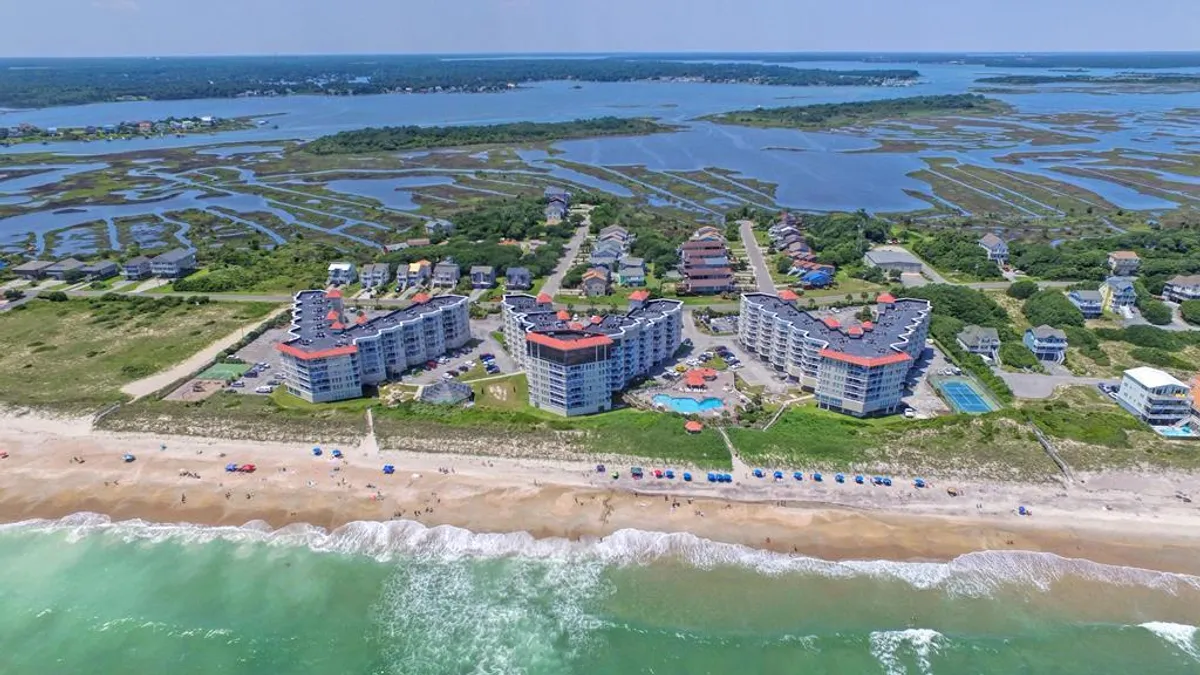 The resort's oceanfront condominium buildings are shown alongside a sparkling swimming pool, with expansive beachfront and surrounding marshland in view. The scene is lively, with areas for swimming, sunbathing, and beach activities, capturing the full scope of the resort's active outdoor amenities.