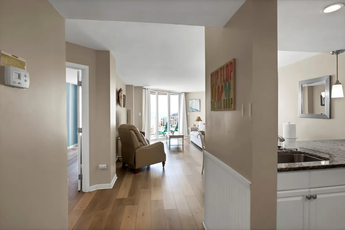 This cozy view includes a kitchen area partially visible in the background, a comfortable armchair by the window, and decorative wall art. The scene suggests an open, multifunctional space where cooking and socializing blend seamlessly.